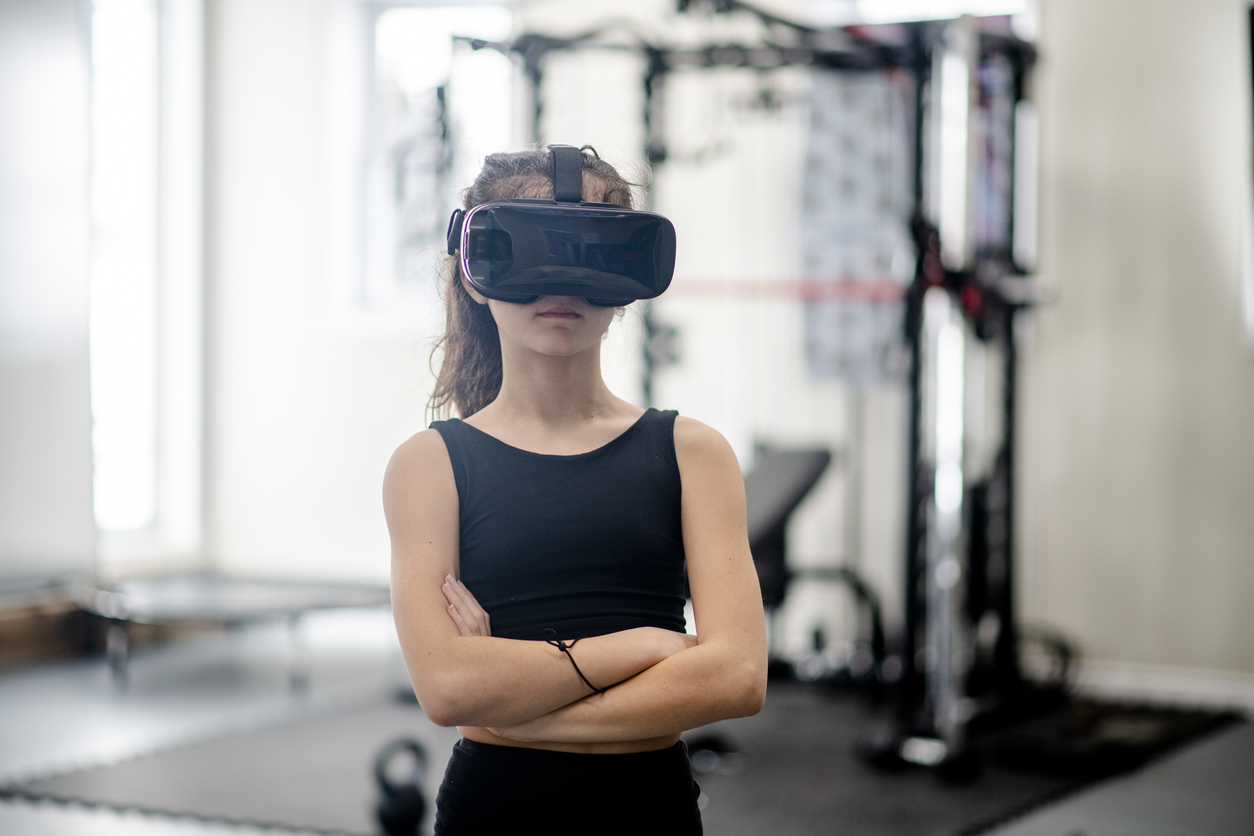 young white woman stands in gym with a VR headset arms crossed